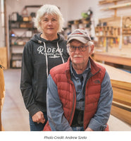 Joan Barrett and Ted Moores pose for a portrait against the backdrop of the Bear Mountain Boats workshop. A caption underneath reads "Photo Credit: Andrew Szeto"