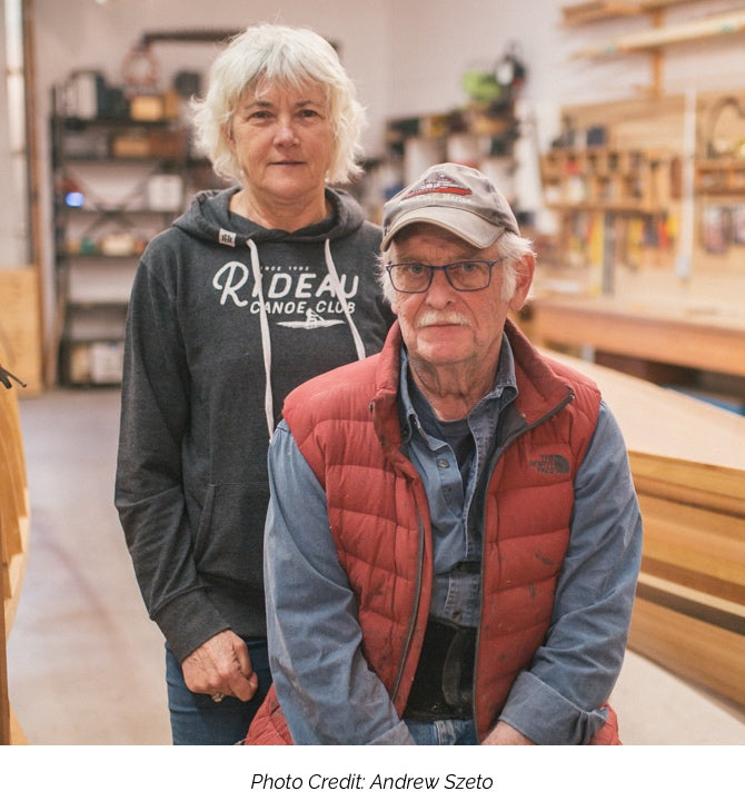 Joan Barrett and Ted Moores pose for a portrait against the backdrop of the Bear Mountain Boats workshop. A caption underneath reads "Photo Credit: Andrew Szeto"